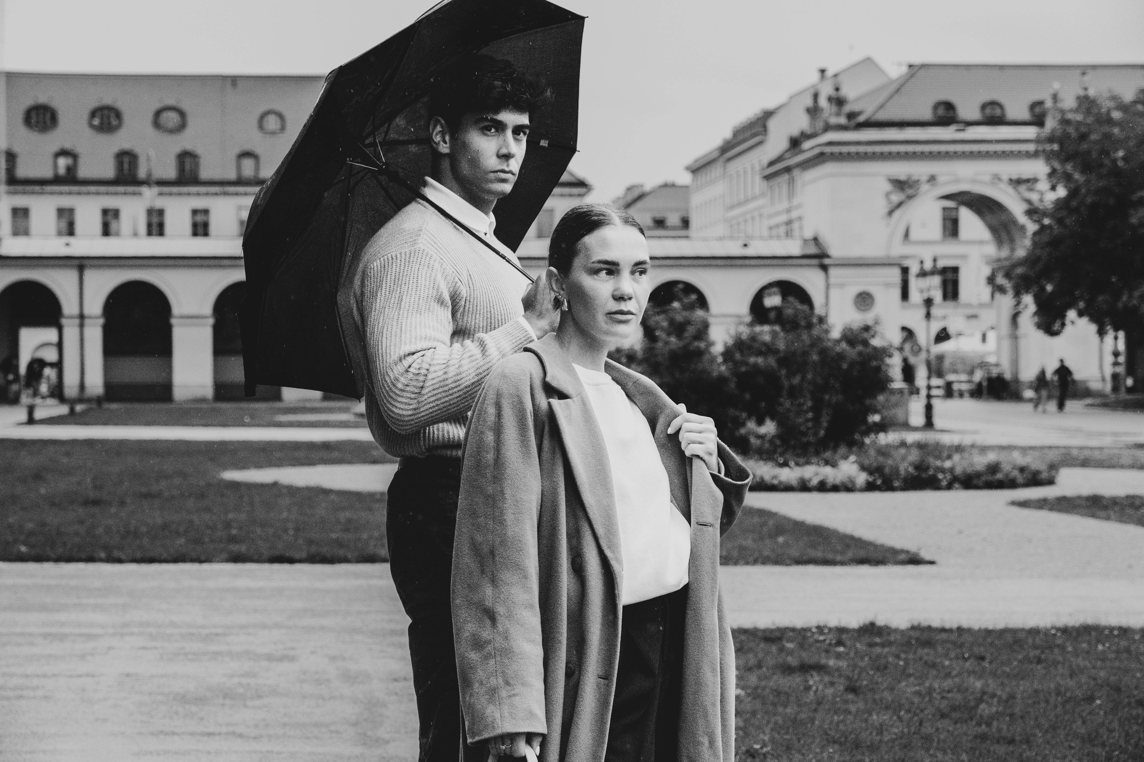 A stylish couple, one holding a sophisticated black Exclusaro luxury umbrella, walking gracefully through a historic European city on a rainy day. Capturing elegance and protection from the elements.
